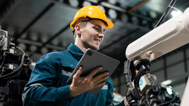 A technician in a blue work uniform and yellow hard hat smiles while holding a tablet, interacting with a robotic arm in an industrial setting. The focus is on the technician's engagement with technology in a manufacturing environment.
