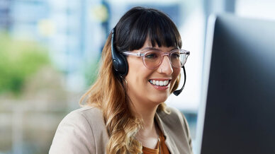 A woman with long, wavy hair and glasses smiles while wearing a headset. She is seated at a desk in front of a computer, engaged in a conversation. The setting appears bright and modern, suggesting a professional work environment.