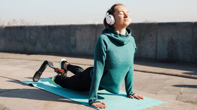 A woman practicing yoga outdoors, wearing headphones and a teal sweatshirt. She is in a cobra pose on a blue mat, smiling peacefully with her eyes closed, enjoying the sunlight. The scene conveys a sense of relaxation and mindfulness.