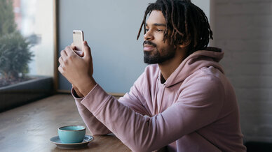 A young man with dreadlocks sits at a café table, smiling while looking at his smartphone. A small blue cup is positioned in front of him on the table, and he gazes thoughtfully out the window, suggesting a moment of relaxation or enjoyment.